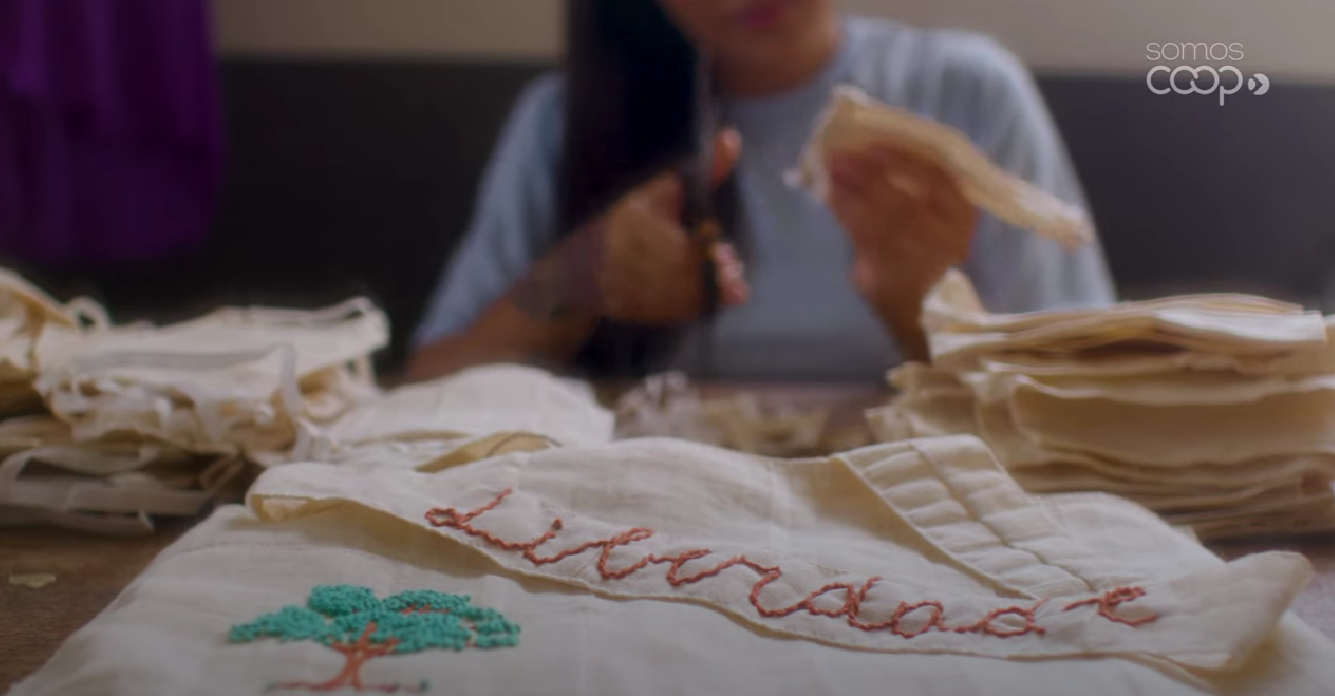 Mulher cortando um tecido ao fundo e em primeiro plano uma bolsa bordada com a palavra liberdade e um ipê.